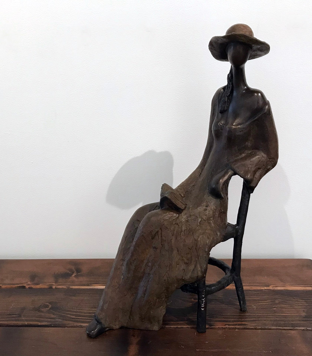 A bronze sculpture of a faceless woman in a long dress and wide-brimmed hat, seated on a wooden chair against a plain background.