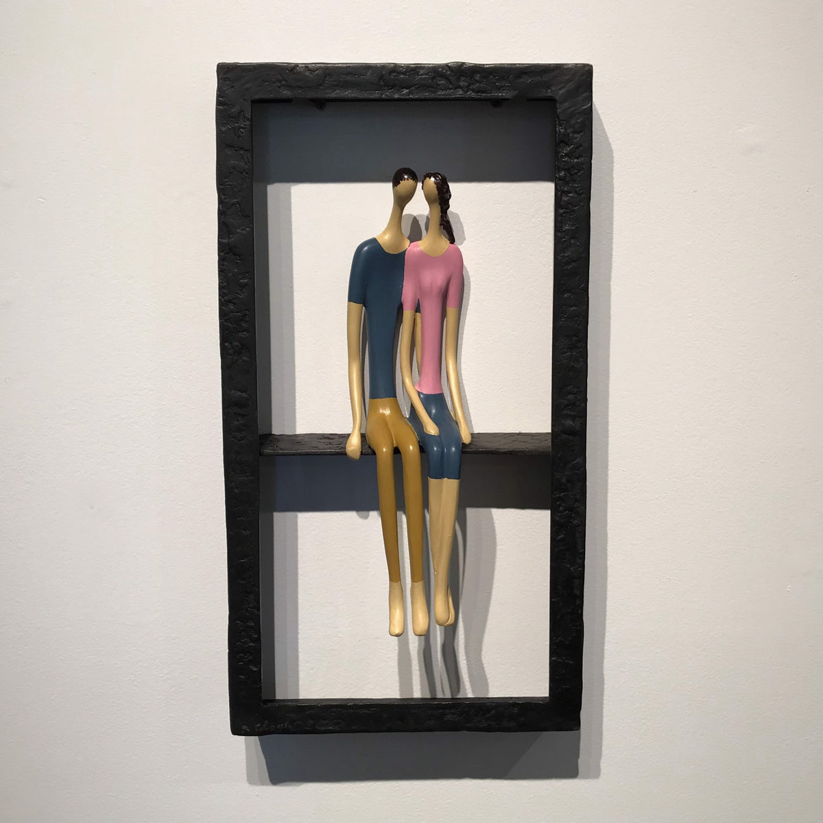 Sculpture of two faceless figures, one in a blue shirt and the other in a pink shirt, sitting together within a black rectangular frame on a white background.