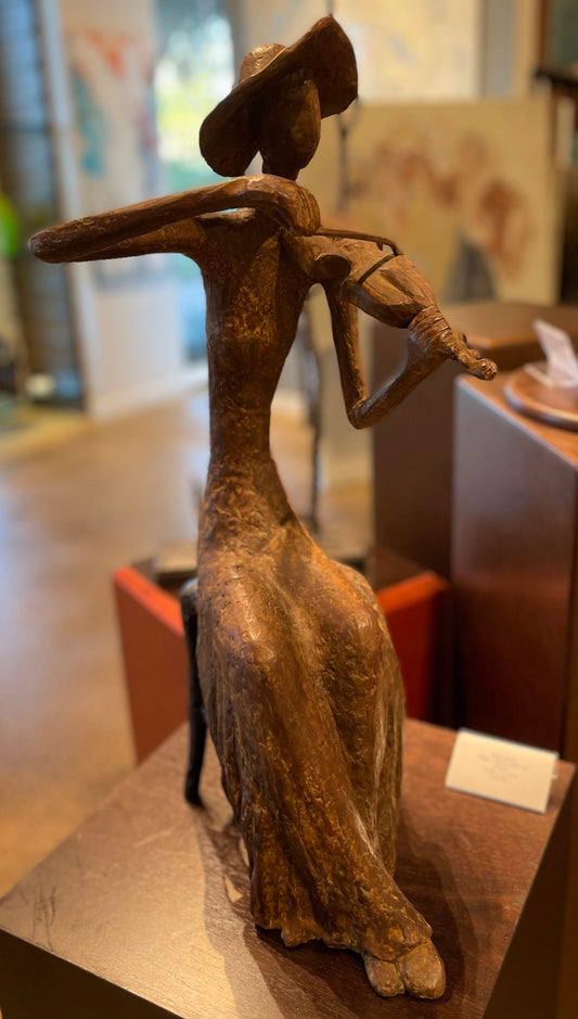 Bronze sculpture of a seated figure wearing a wide-brimmed hat and playing a violin, displayed on a pedestal in an art gallery.