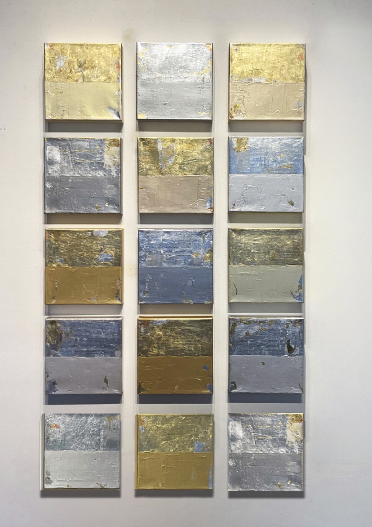 Grid of fifteen abstract panels in gold, silver, and blue tones on a white wall.