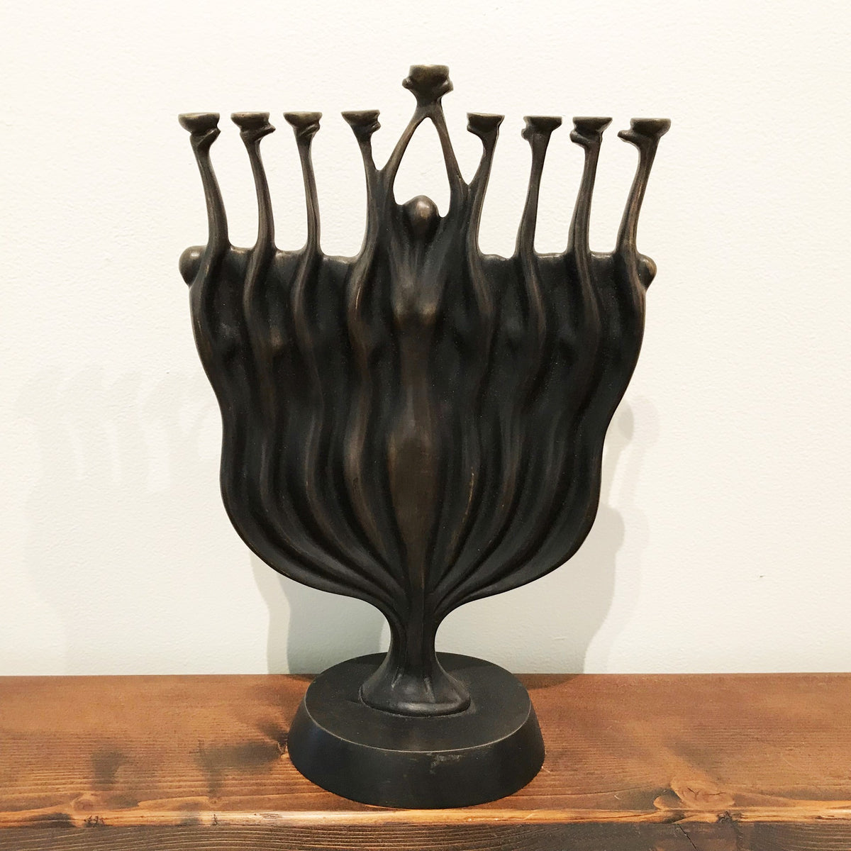 A black statue with multiple elongated figures emerging from a single base turning into a menorah, reaching upwards with their arms extended, displayed on a wooden surface.