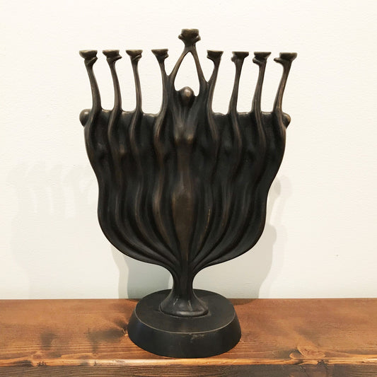 A black statue with multiple elongated figures emerging from a single base turning into a menorah, reaching upwards with their arms extended, displayed on a wooden surface.