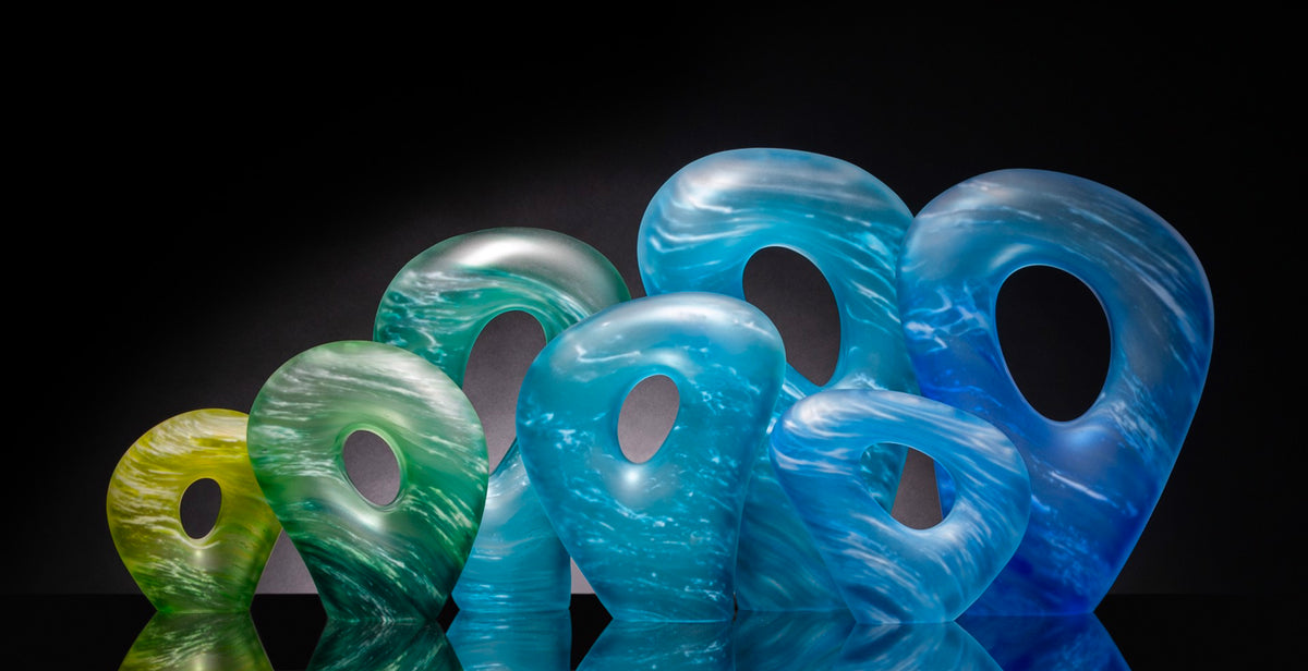 A group of abstract, differently sized glass sculptures in shades of blue and green, each with an oval hole in the center, set against a black background.