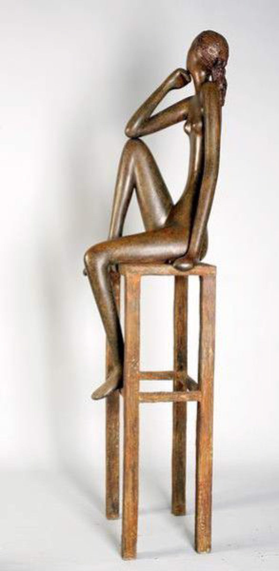 A bronze sculpture of a seated, contemplative figure on a tall, rectangular stool against a plain background.