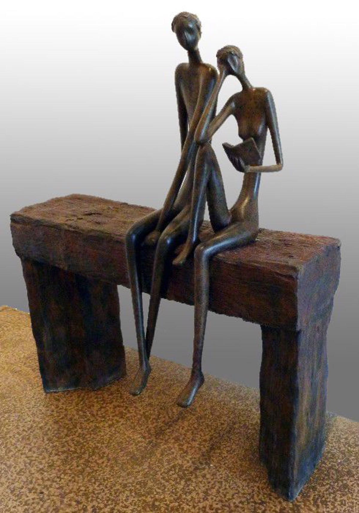 Bronze sculpture of two slender, stylized figures sitting closely on a rustic wooden bench with an abstract, elongated design.