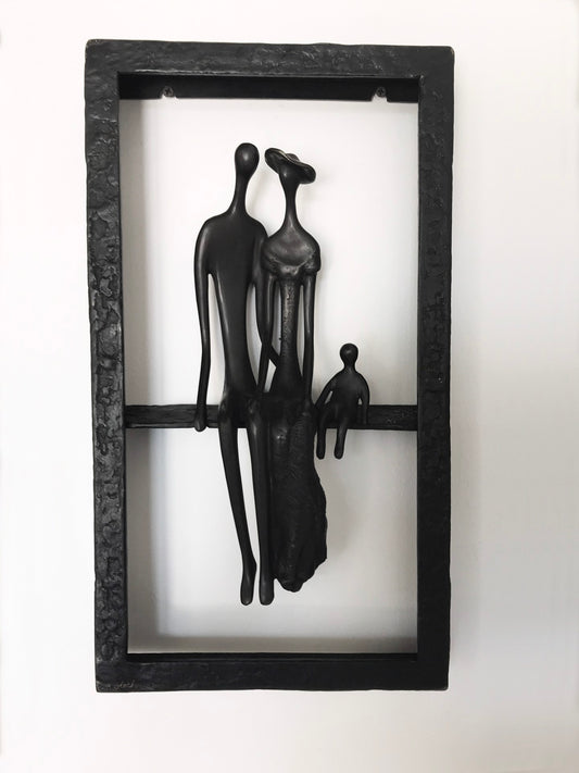A black rectangular sculpture depicts three abstract human figures: a man, woman and child. Mounted within a double-sided frame.