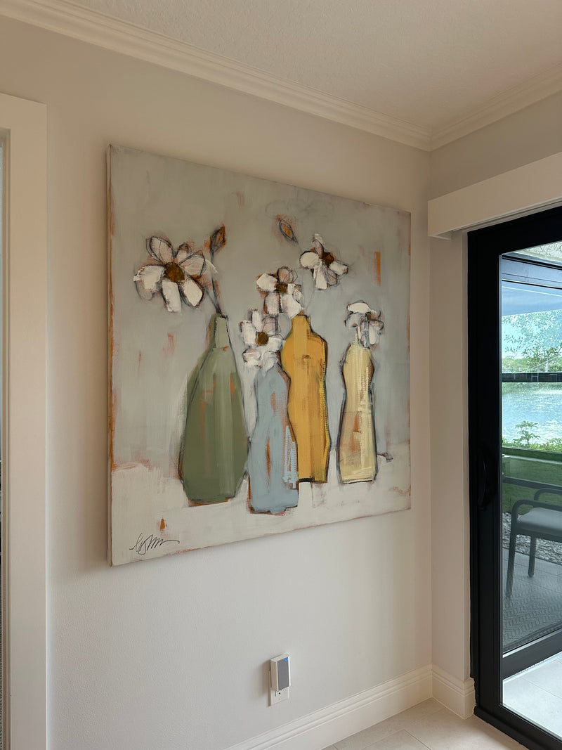 Lynn Johnson painting of white daisies in green, blue and yellow bottles beside a sliding door to a lanai
