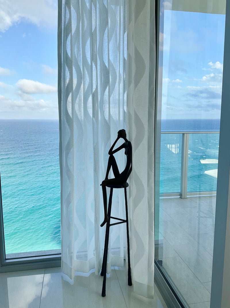 Ruth Bloch bronze sculpture of a seated figure on a tall stool by a window overlooking the Atlantic Ocean