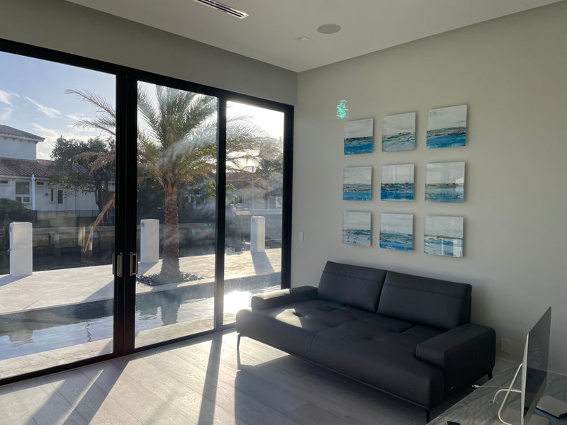 Nine small John Schuyler blue seascape paintings in a grid in a waterfront home with palm tree views
