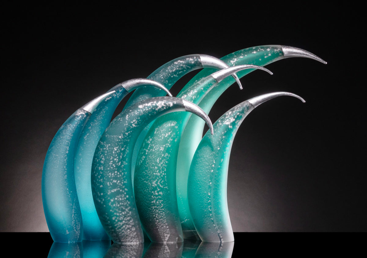 Seven curved teal and green glass sculptures with silver tips on a dark background.