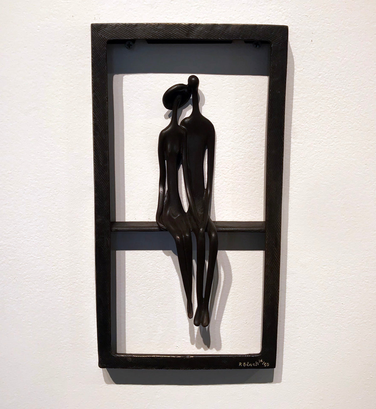 A minimalist sculpture of two abstract human figures sitting side by side within a rectangular frame against a white wall.
