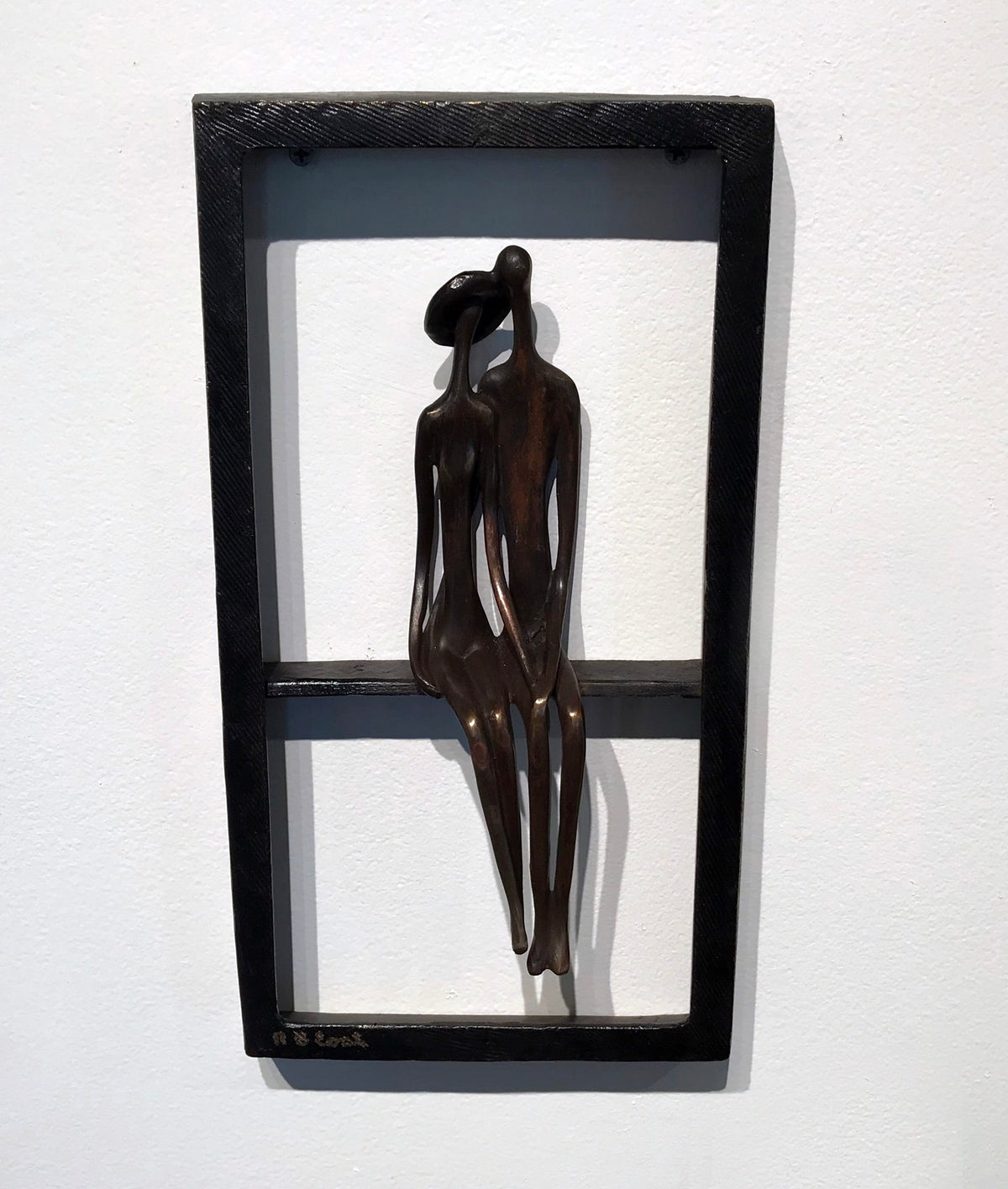 A metallic sculpture of two abstract human figures sitting closely together within a rectangular frame, mounted on a white wall.